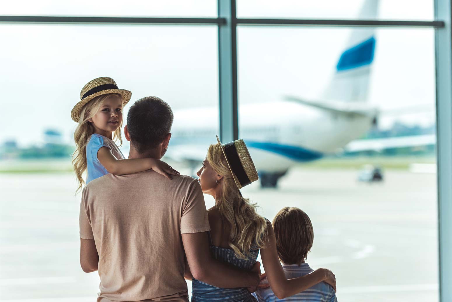 family of four at the airport looking out a window at a plane. Feature image for 9 Tips to Travel More and Spend Less