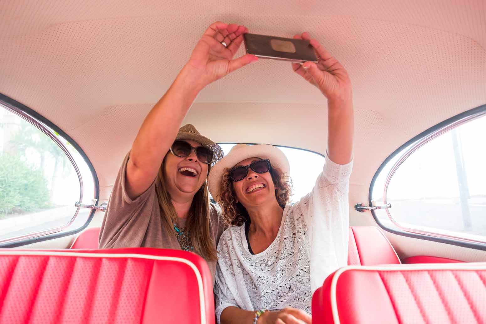 two middle aged women wearing sunglasses taking a selfie in the backseat of a vintage car