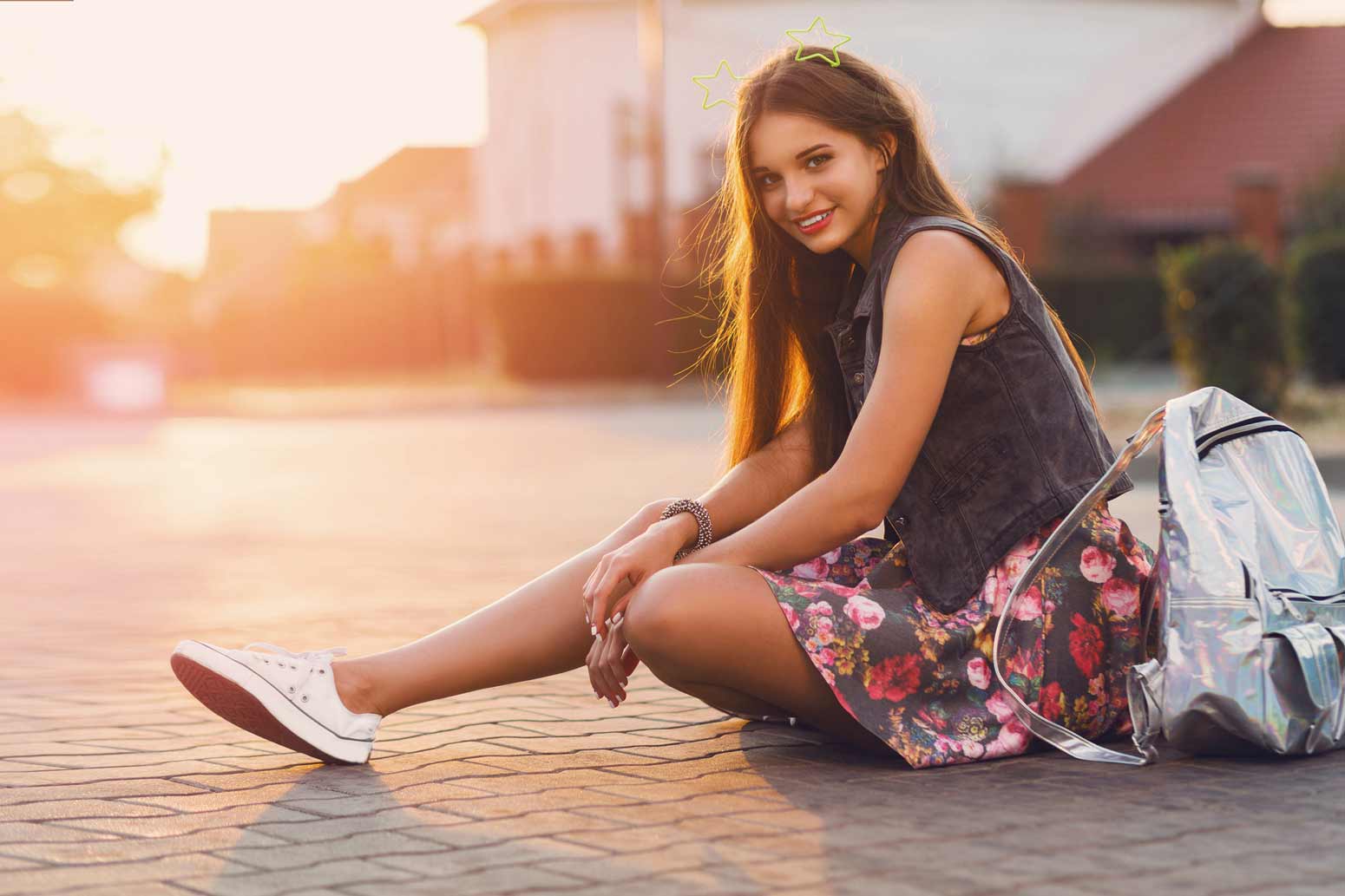 Teen girl wearing jean vest and flower skirt sitting on the pavement outside a building at daybreak smiling at the camera