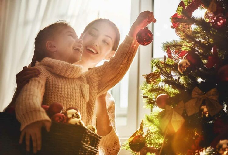 mom helping little girl put christmas tree ornaments on their tree