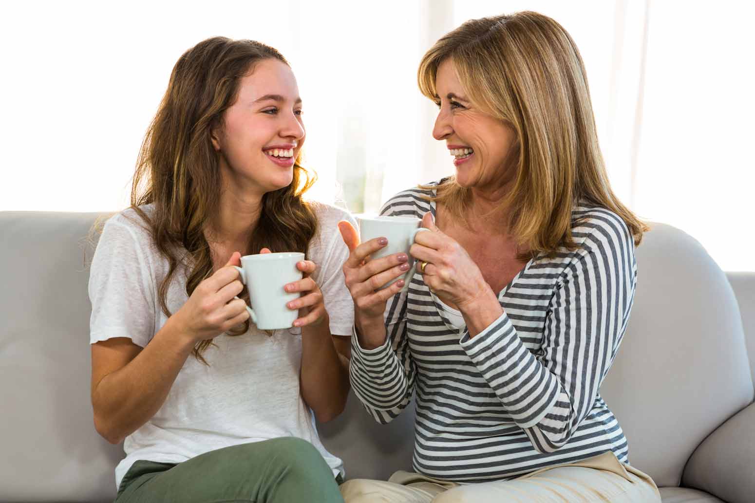 mother-in-law and daughter sitting next to eachother on the couch drinking coffee and smiling and trying to build peaceful relationships