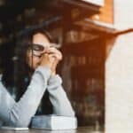 woman sitting at a cafe table with a Bible on it her eyes closed and hands together in prayer because her life feels brittle