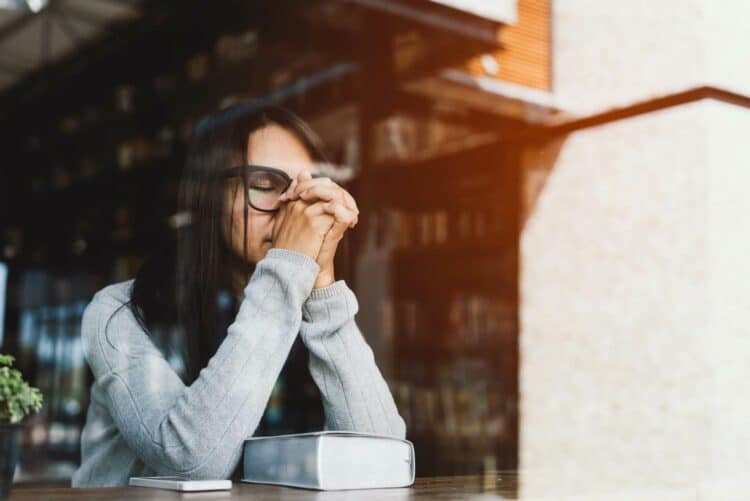 woman sitting at a cafe table with a Bible on it her eyes closed and hands together in prayer because her life feels brittle