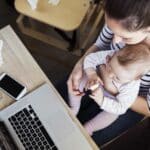 mom holding infant in her lap while working at her computer and wondering "who am I?"