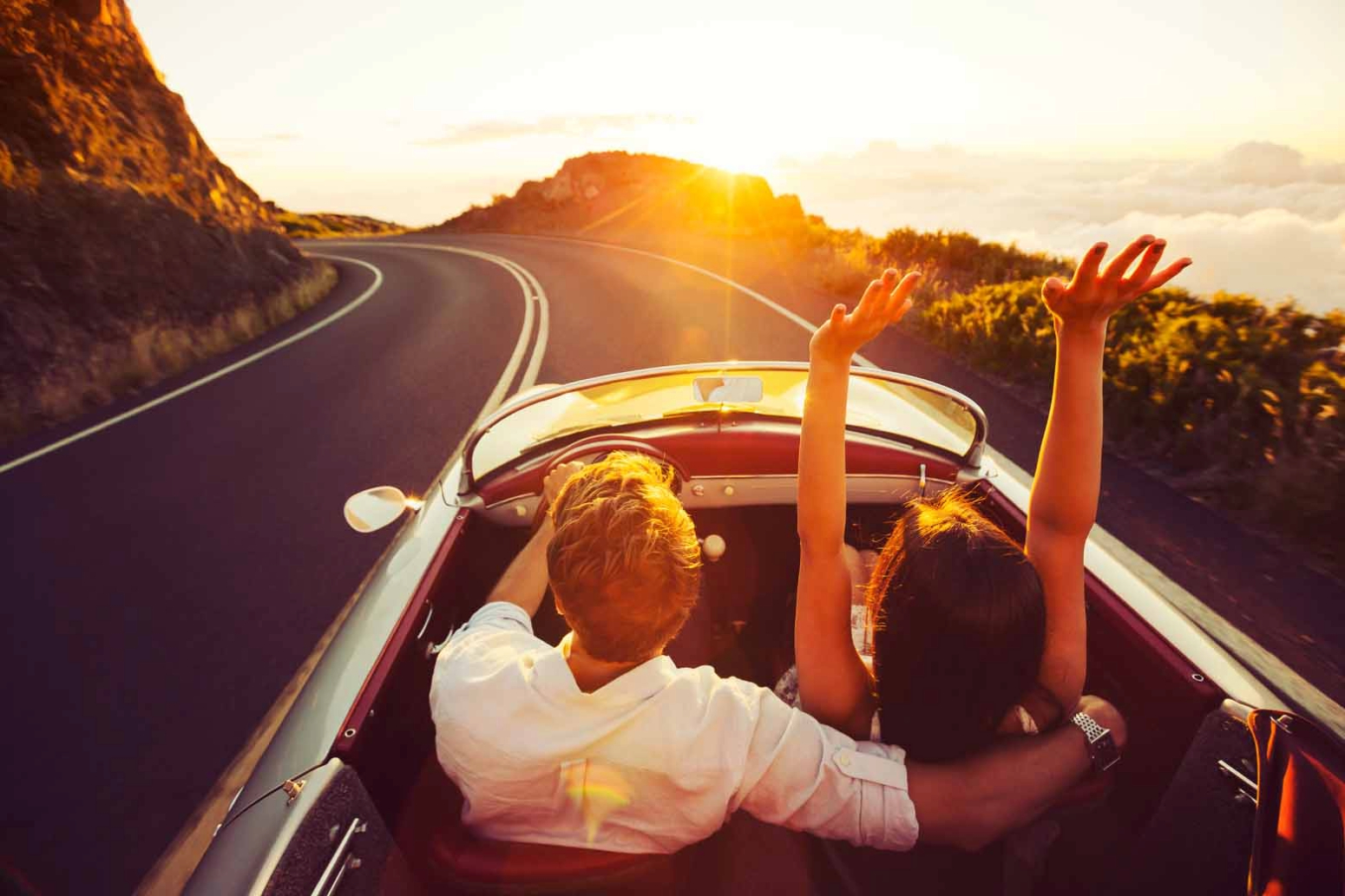 a husband and wife driving along a highway in a convertible at sunset