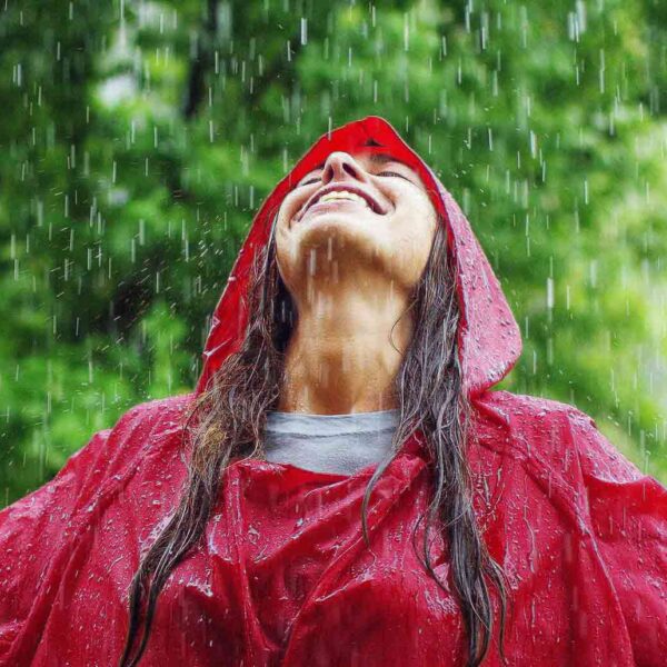 A young woman wearing a red rain jacket practices chasing glimmers by leaning her head back and laughing in the rain