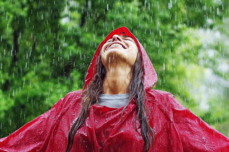 A young woman wearing a red rain jacket practices chasing glimmers by leaning her head back and laughing in the rain