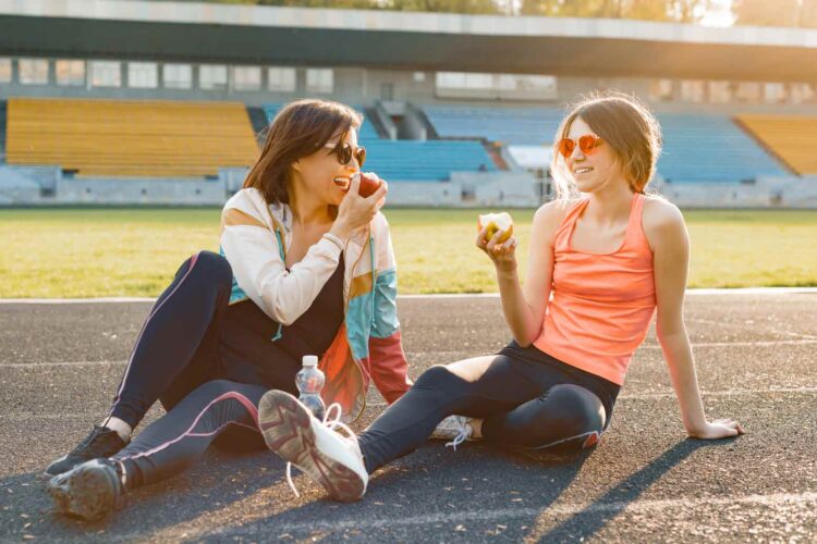 mom sitting on a race track eating apples and laughing with her daughter while getting her teen ready to launch