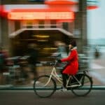 women wearing a red coat riding a bike in a city block and speeding through life