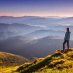 a woman stands at the edge of a cliff overlooking a smoky blue mountain range contemplating whether you are living your one life well