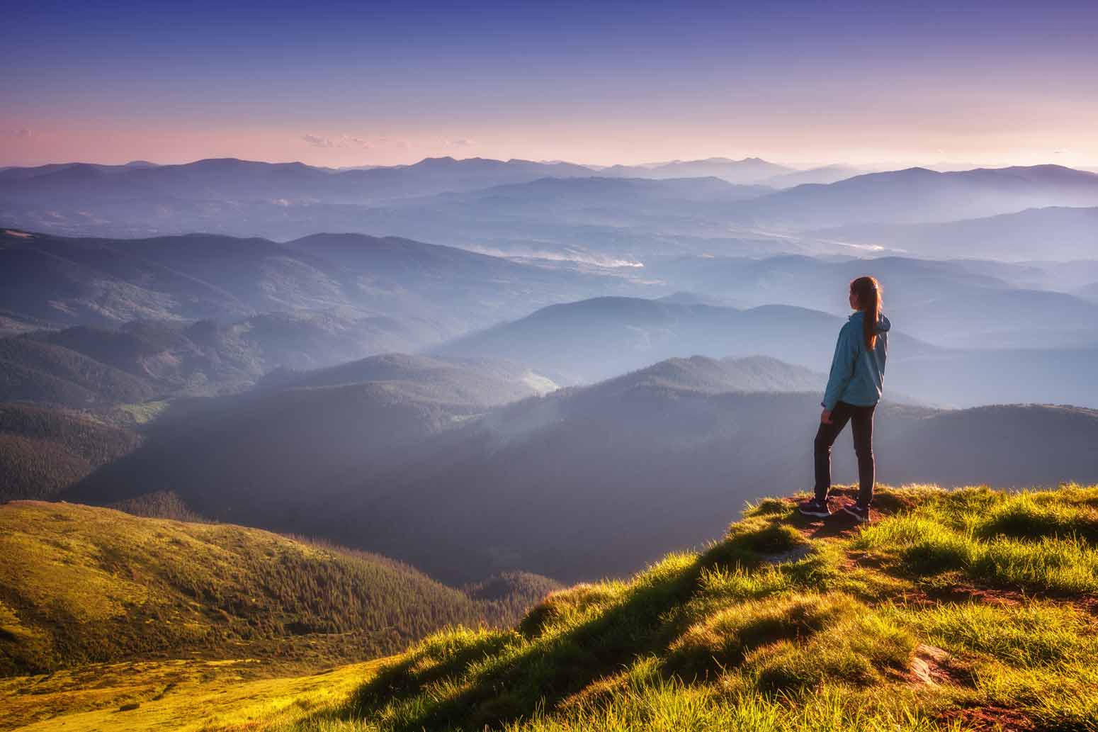 a woman stands at the edge of a cliff overlooking a smoky blue mountain range contemplating whether you are living your one life well
