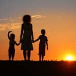 silhouette of a mom holding hands with her two young kids in front of a setting sun, a symbol of making them a lifetime promise of her love
