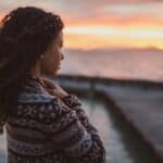 a woman with a serious expression looks out over the water at sunset wondering "Where is God when it hurts?"