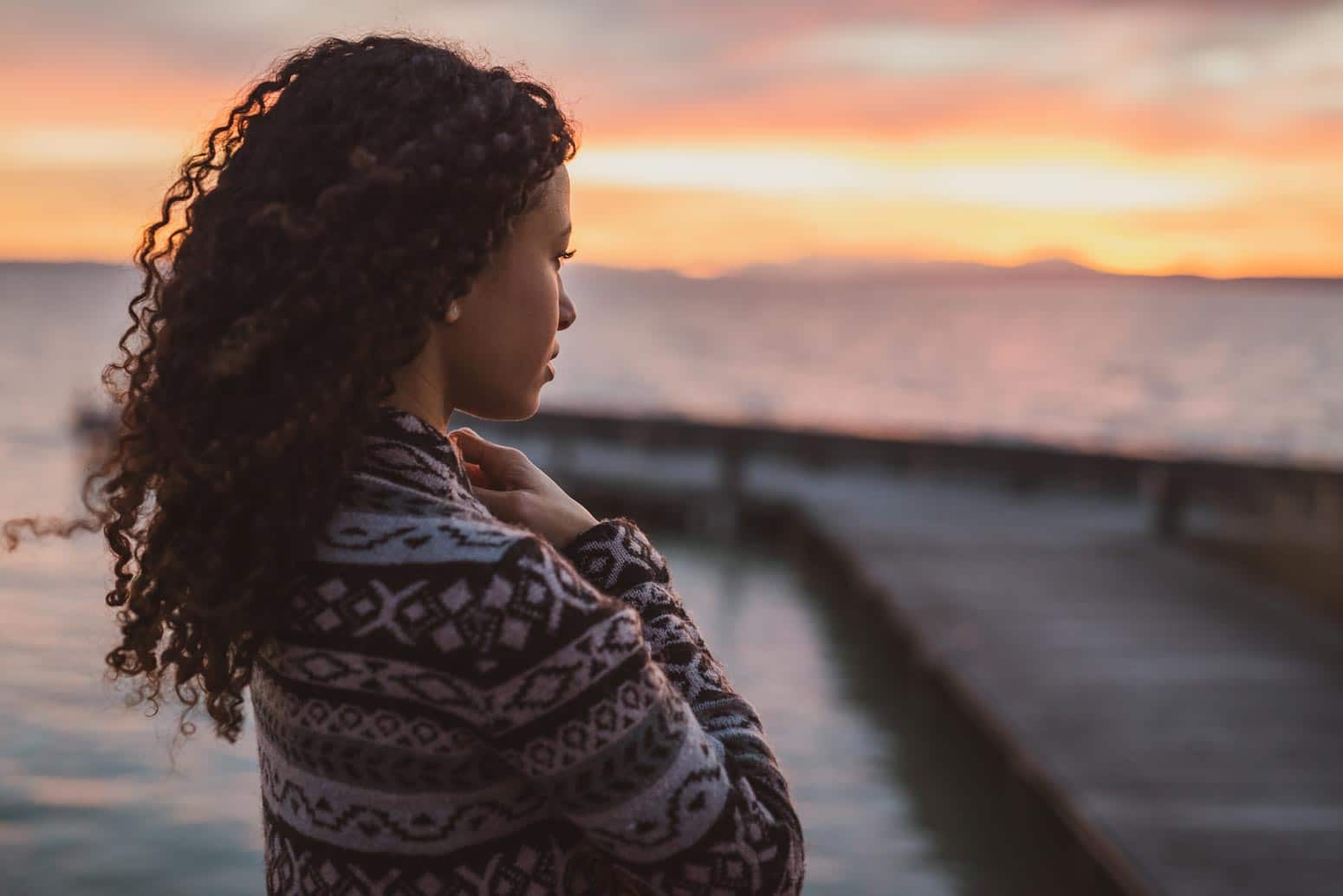 a woman with a serious expression looks out over the water at sunset wondering "Where is God when it hurts?"