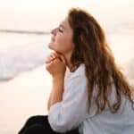 a woman sits on the beach with her eyes closed and hands folded under her chin, wondering how to know God is speaking to you
