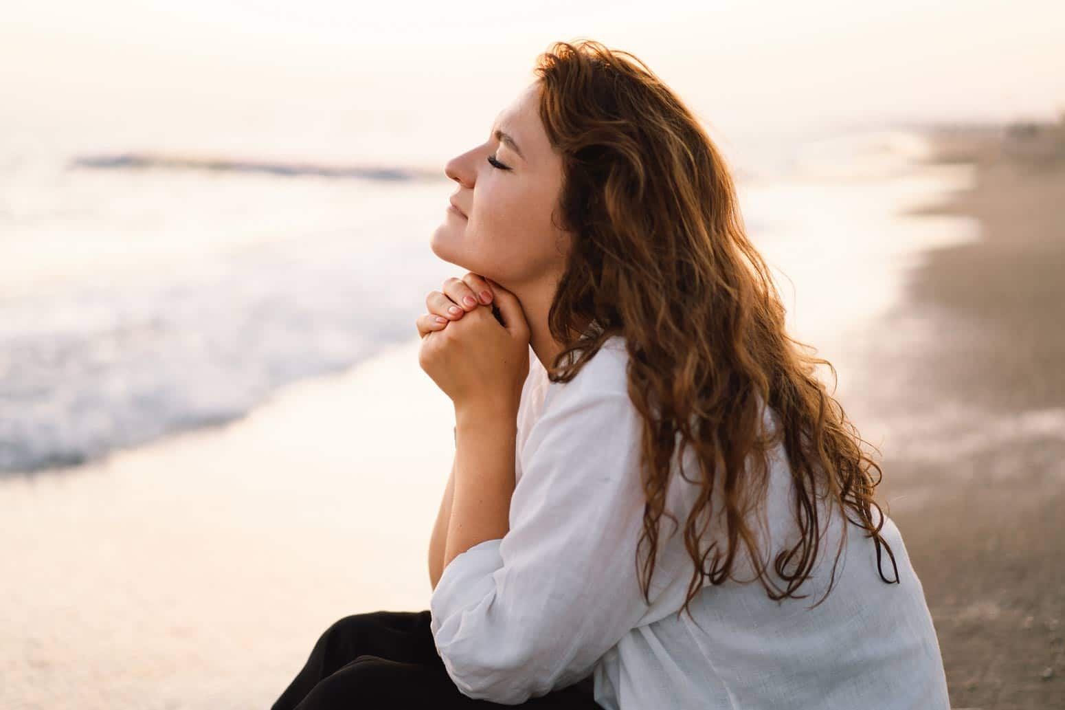 a woman sits on the beach with her eyes closed and hands folded under her chin, wondering how to know God is speaking to you