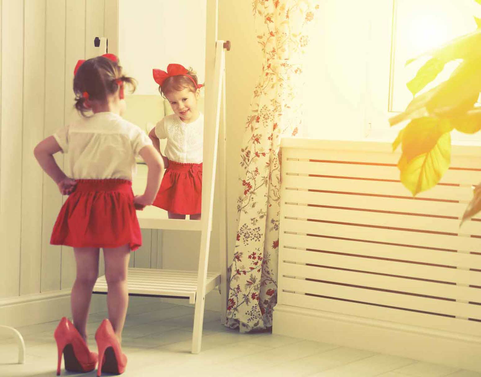 a little girl in her mom's heels studies herself in a mirror, underscoring the importance of protecting your child's self-worth