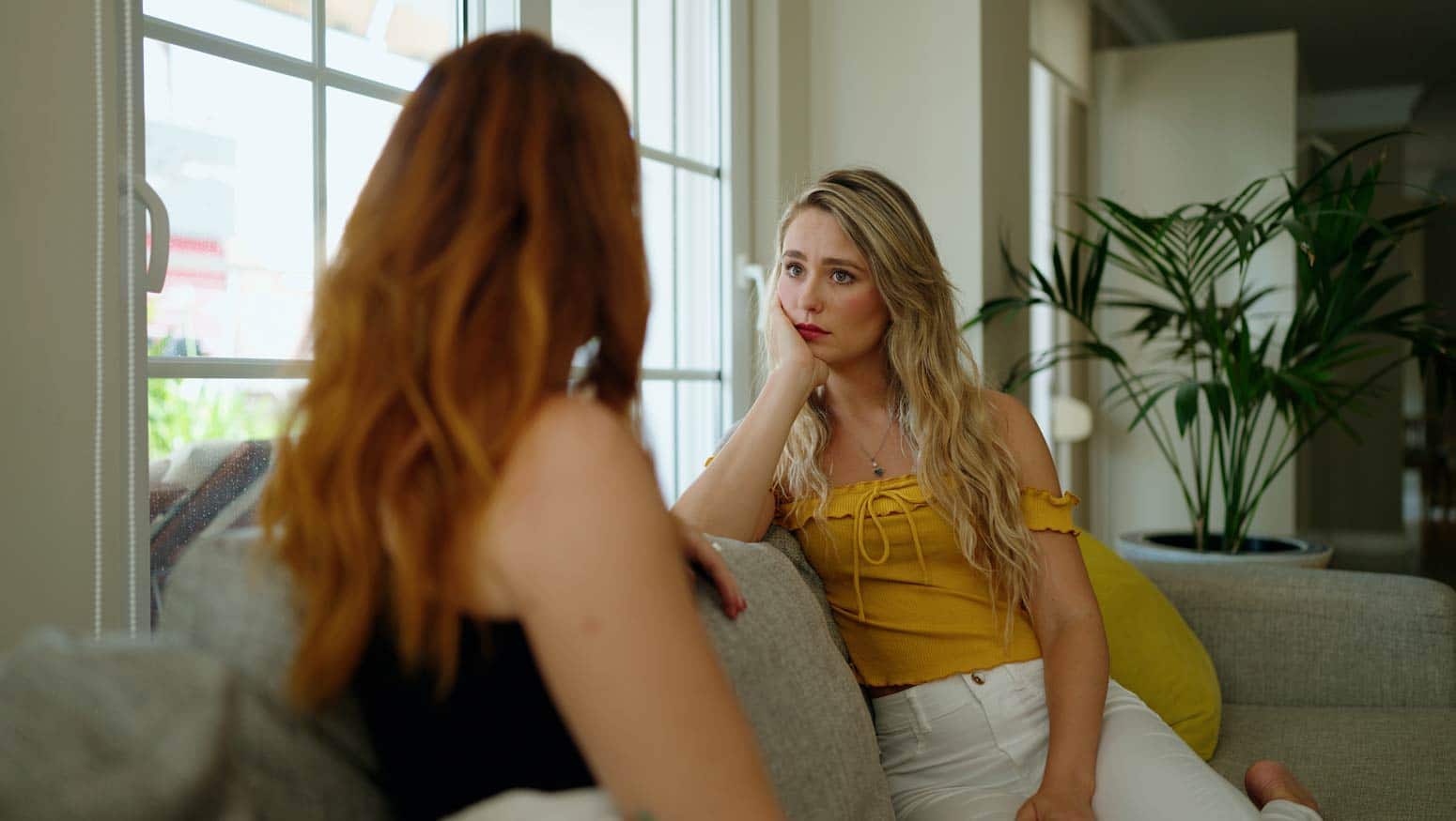 two young women sit on a couch facing each other with a grim expression on their face as they contemplate how to restore a broken relationship