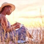woman sits near the ocean at sunset writing in a journal as a healing act of self-care