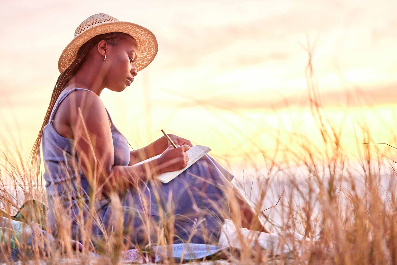 woman sits near the ocean at sunset writing in a journal as a healing act of self-care