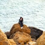 woman sits on a rocky cliff near the water looking thoughtful and anchored in faith