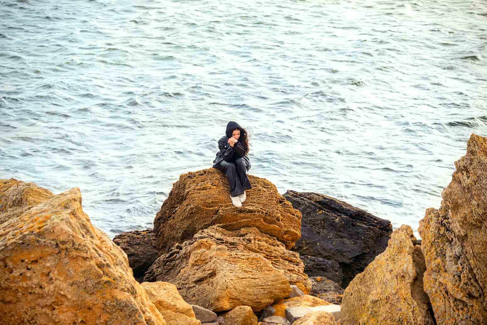 woman sits on a rocky cliff near the water looking thoughtful and anchored in faith