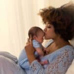 a woman sitting up on a bed kissing her newborn baby on the head and leaning into her new identity as a mom