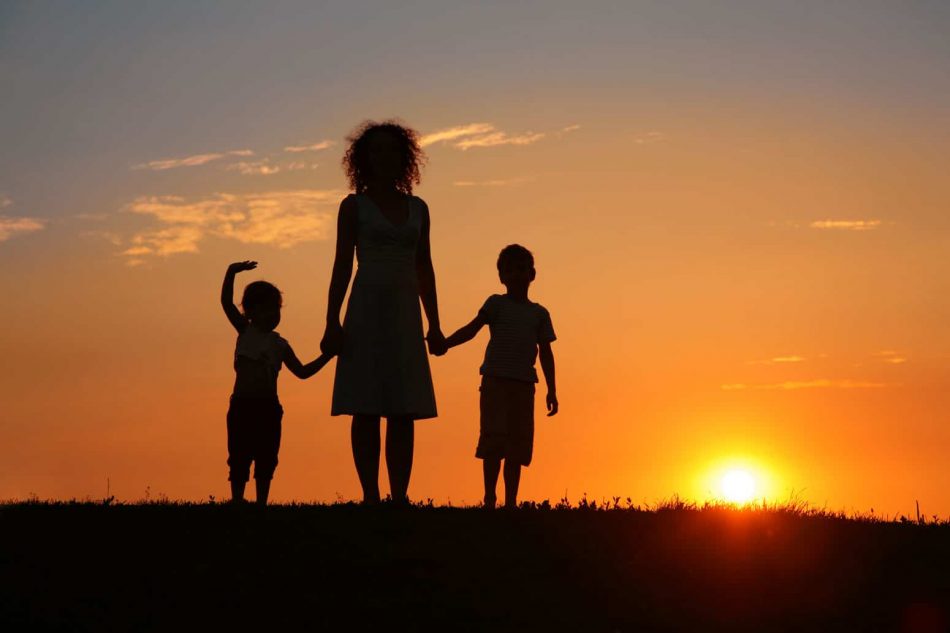 silhouette of a mom holding hands with her two young kids in front of a setting sun, a symbol of making them a lifetime promise of her love